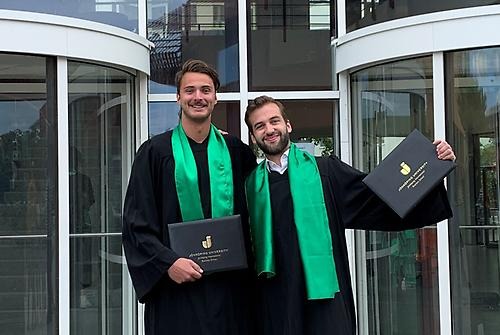 Sören Pontgratz and Niklas Koch in their graduation gowns facing the camera, holding their degree diplomas up with one hand and arms around each other's shoulder. They are standing in front of JIBS entrance. 