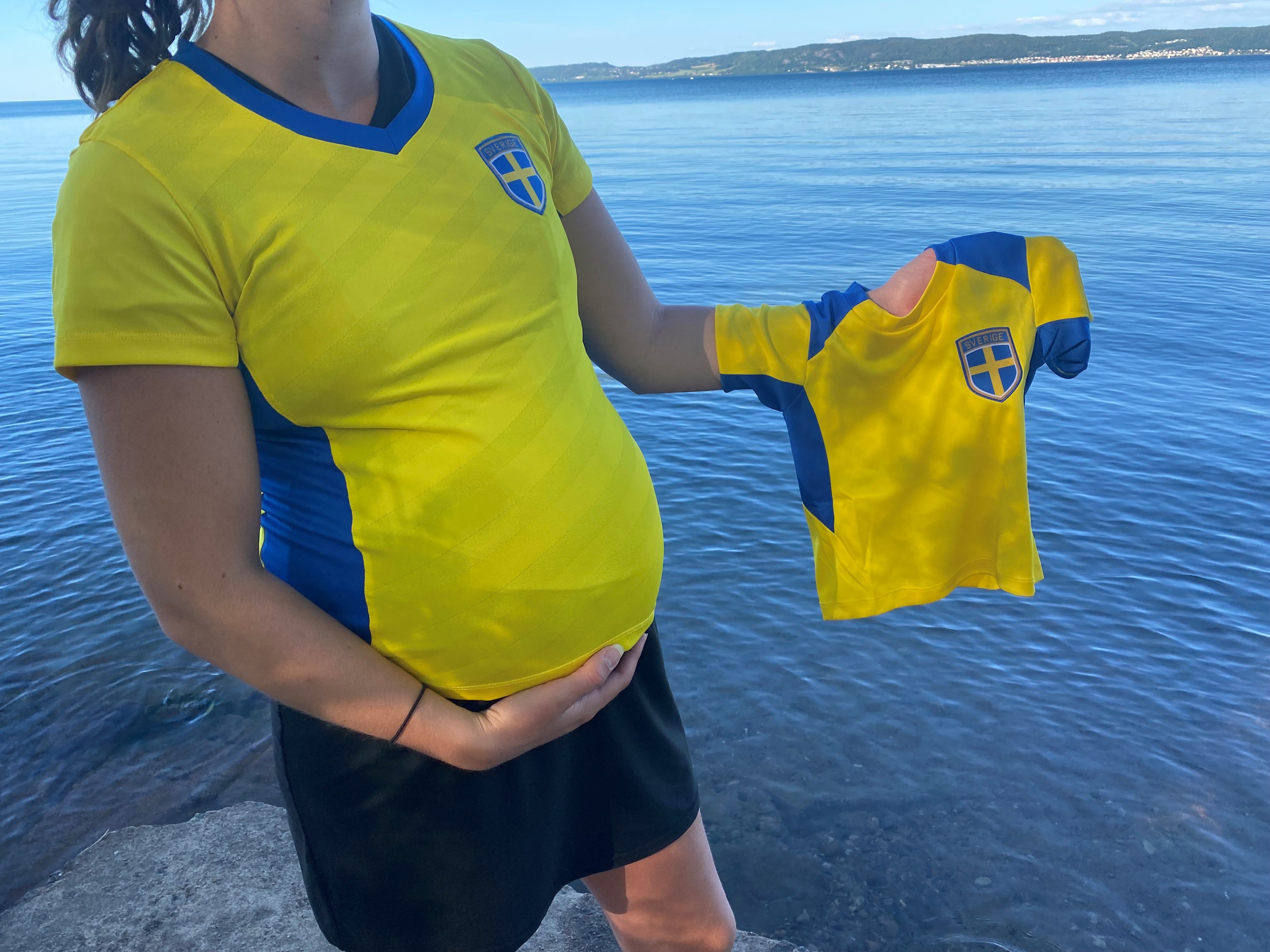 Gravid kvinna som har på sig en fotbollströja och håller upp en fotbollströja som passar för bebisar.